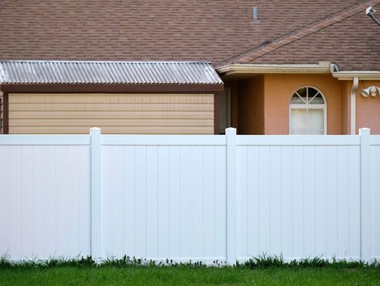 A white PVC fence installed in front of a peach-colored home with a metal shed in its yard during the daytime.