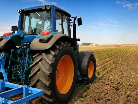 Blue tractor working on a cultivated field with rows of crops under a clear blue sky in a rural farming landscape.