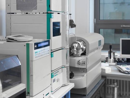 A laboratory interior with several white and teal machines on a countertop near a desk with a monitor and keyboard.