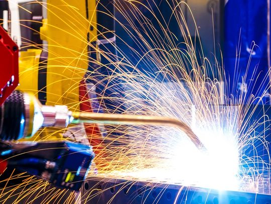A yellow robotic arm welds a rectangular metal part, emitting yellow-orange sparks from the point of contact.