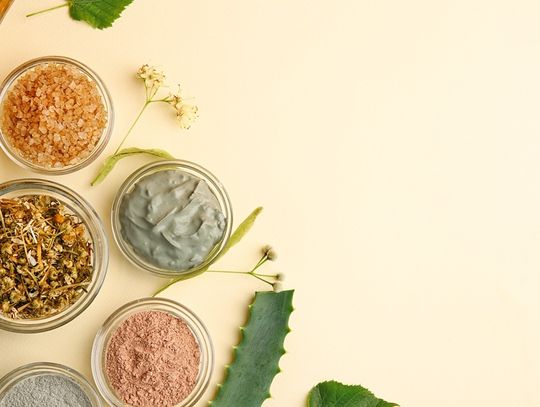 Top view of cosmetic ingredients like clay masks, dried herbs, bath salts, and oil arranged on neutral surface.