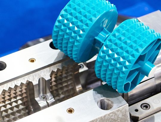 A finished, cyan plastic part with two spike-covered cylinders sitting on top of its silver metal mold.
