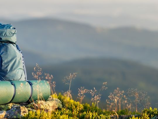 A blue hiking backpack and rolled sleeping pad resting on rocks and grass with blurred mountains in the background.