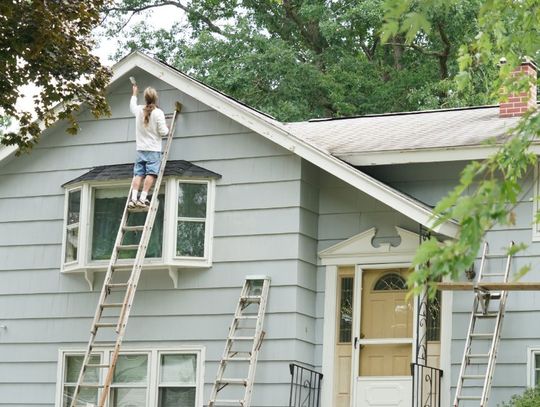 Person on a ladder paints the upper siding of a gray house, with extra ladders and trees around the yard.