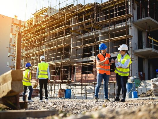 Construction workers stand around an active build site with safety vests and hard hats on. Two are talking about plans.