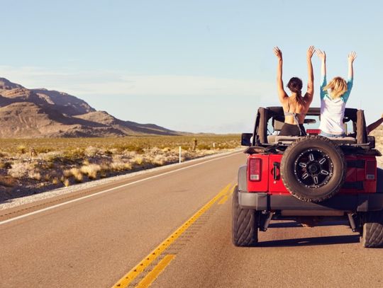 A red vehicle driving on a desert road with passengers raising their arms near mountains under a clear sky.