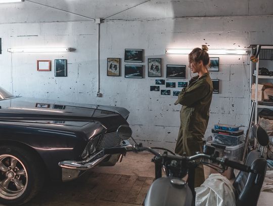A blonde woman in a mechanic suit stands with her arms crossed in front of a vintage car parked in a garage.