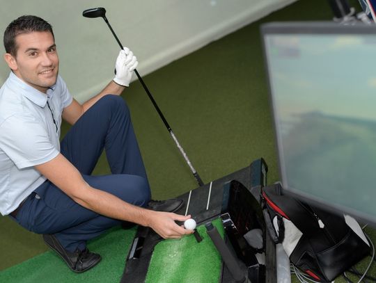 A man smiles as he positions a small golf ball on his simulator. He holds a golf club in his other hand.