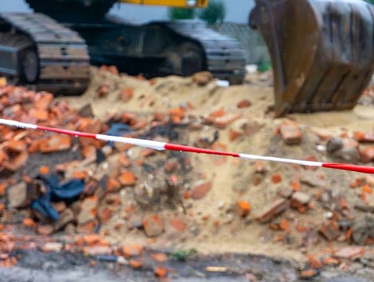 A crawler excavator is poised above a mound of dirt and bricks behind a line of red-and-white warning tape.