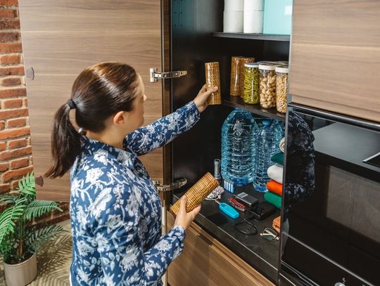 A woman organizes a modern kitchen pantry with jars, bottled water, and emergency tools, set against a brick wall.