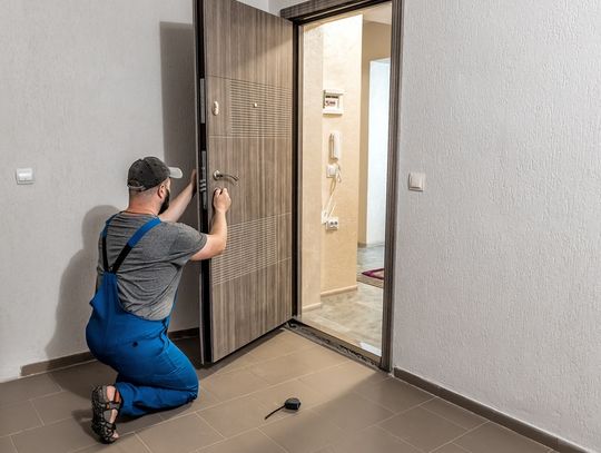 A person wearing a hat kneeling and installing a large beige front door in the entryway of a modern home.