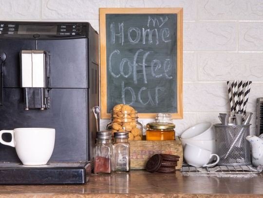 A bar has an espresso machine with a white mug on it. Different coffee ingredients are near, and a chalk sign says "my home coffee bar."