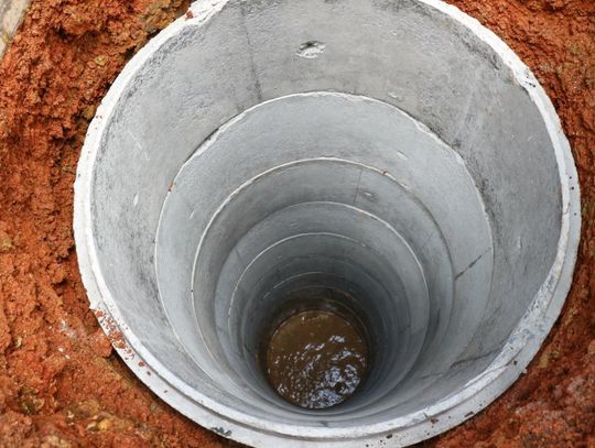 A bird's eye view shows a concrete well filling up with water and surrounded by orange clay mud.