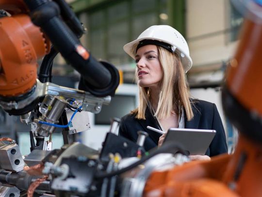 A professional woman wearing a white hard hat holds a tablet while she examines the components of a machine.