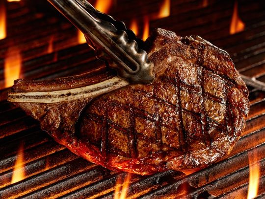 Tongs lift a Bone-In Ribeye steak as it grills over open flames, showing perfect char marks and a caramelized crust.