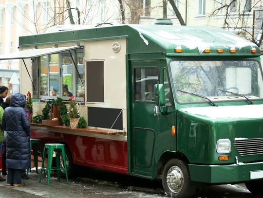 A green food truck parked outside during the daytime during winter. Three customers wait outside the truck.