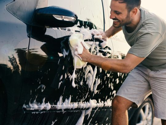 A close-up of a man smiling while using a sponge with soapy water to clean the exterior of a black vehicle.