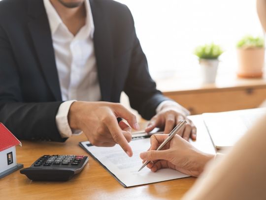 A close-up of a hand holding a pen over a document on a desk in front of a man pointing at a line on the document.
