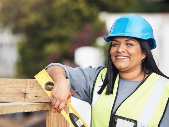 A construction worker wearing a safety vest and a hard hat leaning on a wooden structure with a level in her hand.