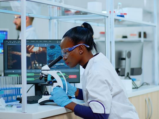 A young Black female scientist checking sample of virus using microscope in modern lab. She's wearing safety gear.