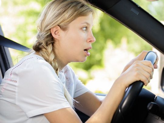 A blonde woman looks shocked as she grips her car's steering wheel and stares wide-eyed through the windshield.
