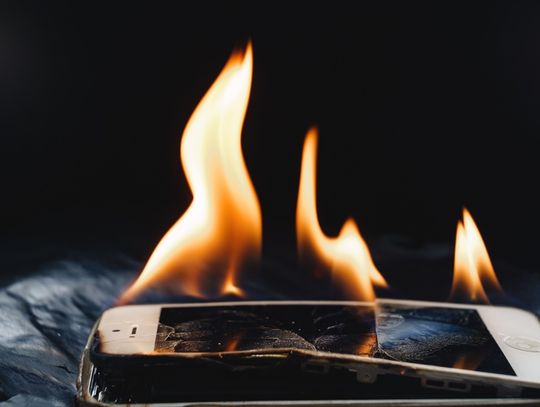 A cell phone on fire with a cracked, bent screen with smaller flames. The screen is splitting on the edge.