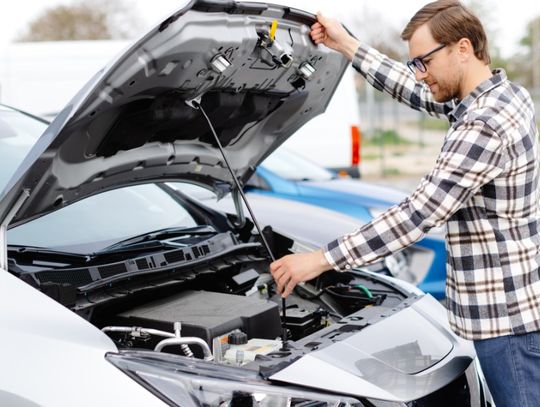 A person wearing glasses opening the hood of a gray car parked outside next to a blue car during the daytime.
