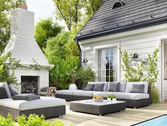 Luxury backyard patio with modern outdoor seating, a white brick fireplace, lush landscaping, and an open view into a stylish kitchen.