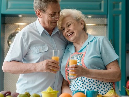 A senior couple is leaning against each other while holding cups of juice. A counter with produce is in front of them.