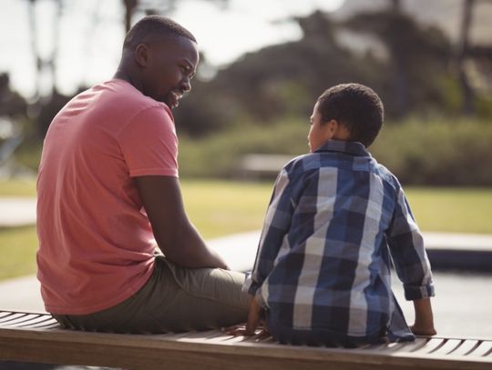 A man sits on a wooden bench next to an elementary school-aged boy at a park. They are facing each other.