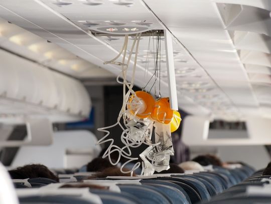 Several yellow oxygen masks with white tubing drop from the aircraft cabin ceiling above seated passengers.