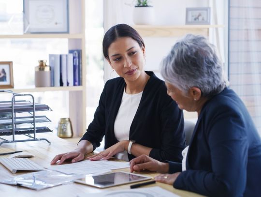 An older woman is sitting next to a younger woman at a desk that has several pieces of paper on it.