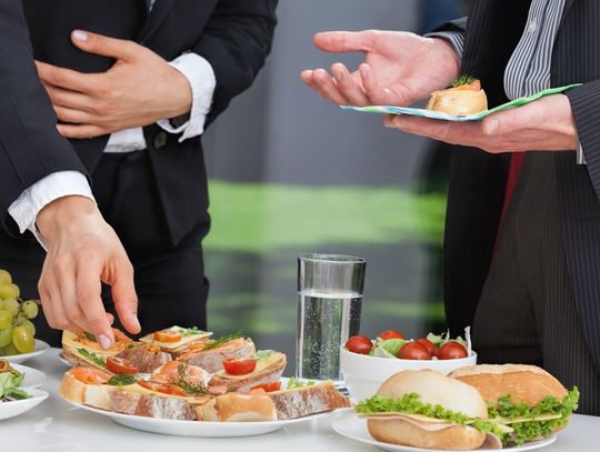 Two business professionals wearing nice suits stand at a lunch buffet in their office. One reaches to pick up a small sandwich.
