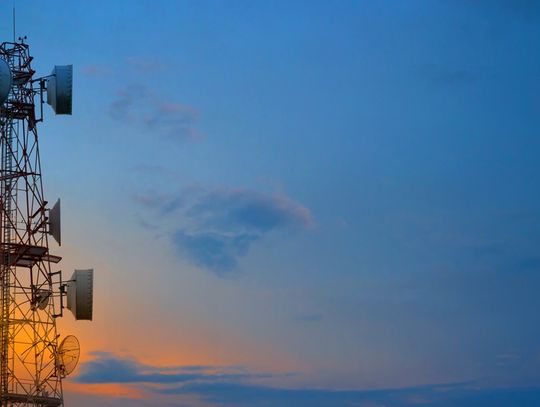Two telecommunication towers with wireless antennas against with the sun setting behind clouds behind them.