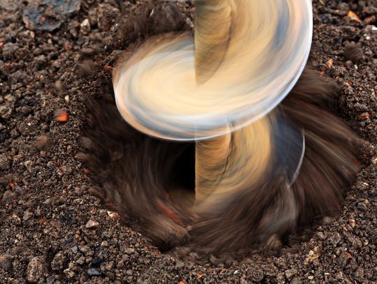Close-up of a rotating earth auger drilling into dark soil, creating a deep hole as loose dirt spins around the bit.