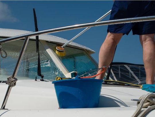 A man scrubbing a white boat using a brush while a pressure water system sprays water with a clear sky in the background.