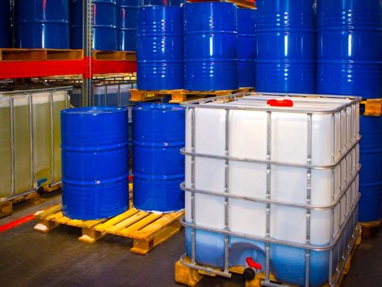 Several blue metal barrels are organized and stacked in an industrial warehouse. A clear, plastic container sits near them.
