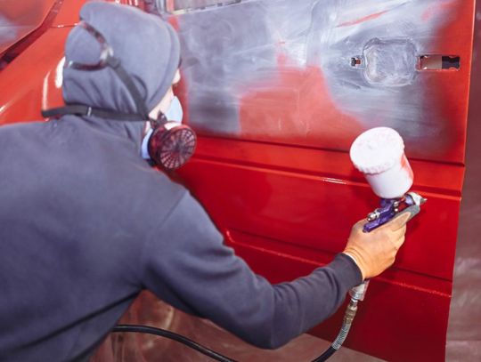 A person wearing a gray hoodie and a respirator mask uses a spray paint gun to paint a damaged car red.