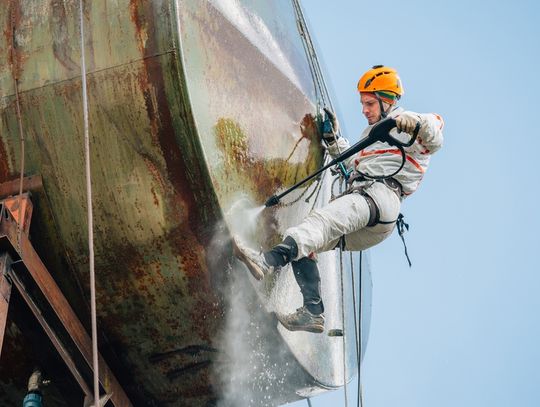A worker in a safety harness using a high-pressure hose to wash the curved side of a large industrial storage tank.