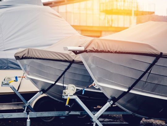 A close-up of a row of boats, each covered with a fabric boat cover. They are lined up in front of a storage building.