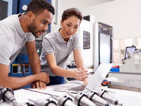 Two engineers wearing gray shirts look at a laptop on a large table in a bright manufacturing facility.