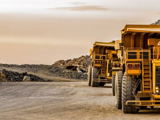 Yellow mining trucks drive down a dirt road in a mining quarry with piles of mined material and debris off to the side.