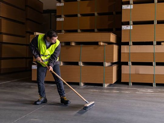 A man wearing a bright green safety vest is using a push broom to clean the floor of a warehouse full of pallets.