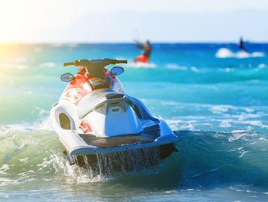 A jet ski floats on top of some water with other people having fun in the background. The sun shines from the left.