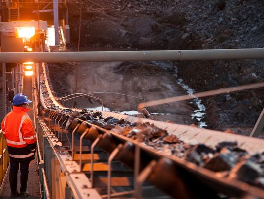 A person wearing an orange jacket and a blue hard hat observes a conveyor belt moving ore rocks in a mine.