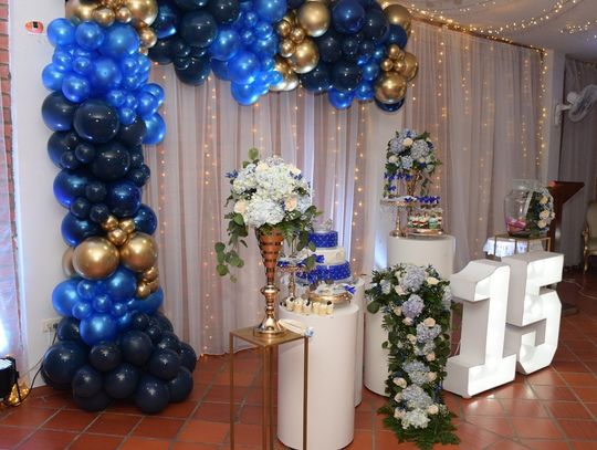 A room with a blue and gold balloon arch, string lights, and pillars with flower bouquets and a tiered cake.