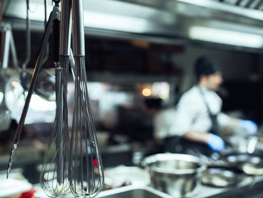 Metal utensils hanging in a commercial kitchen behind a restaurant. A chef is cooking on a stovetop nearby.