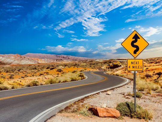Winding road through a rocky desert landscape with a yellow curves warning sign indicating winding road for four miles.