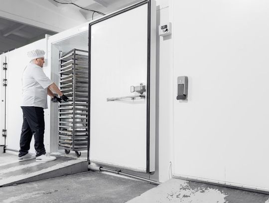 A man wearing a white uniform and a chef's hat, pushing a metal cart with stackable trays inside an industrial freezer.