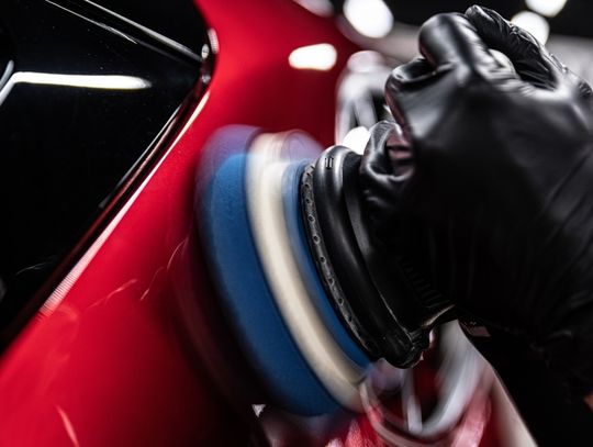 A close-up of a professional using an electric machine for paint correction in a red car detailing studio.